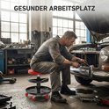 Man in werkpak zit op rode rolkruk naast een auto met open motorkap. Tekst 'GESUNDER ARBEITSPLATZ'.