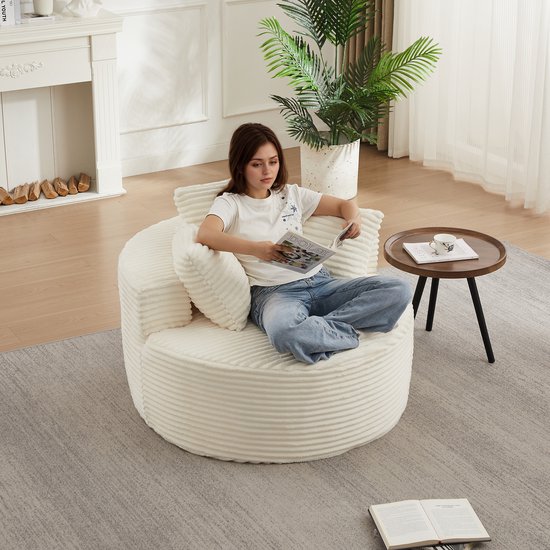 Een jonge vrouw zit op een crèmekleurige corduroy fauteuil met ribbelpatroon en drie kussens, lezend in een tijdschrift.