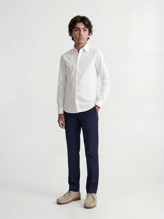 Garçon en chemise blanche à manches longues et pantalon bleu marine, debout.
