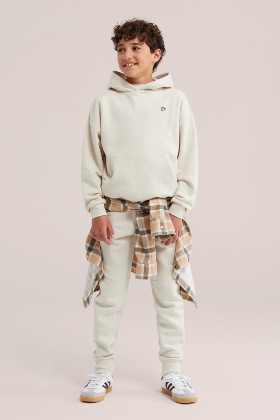 Jongen in een lichtbeige hoodie met geborduurde duif, een geruite blouse om de heupen en bijpassende joggingbroek.