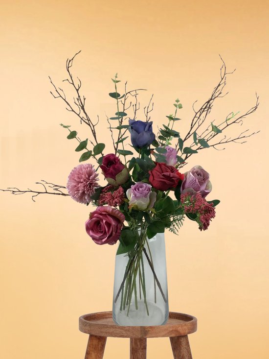 Un bouquet de diverses fleurs en soie de couleurs rose, bordeaux et violet, avec des branches et de l'eucalyptus, dans un vase en verre sur un tabouret en bois.