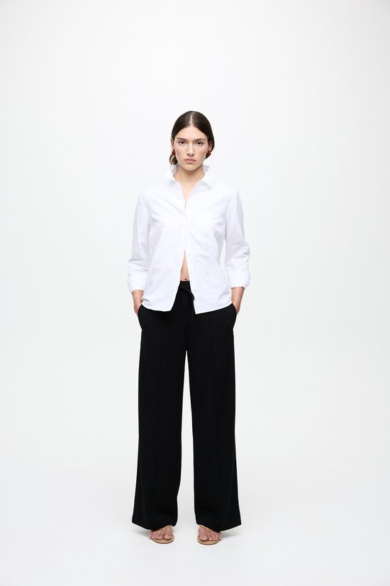 Model met handen in zakken, draagt een zwarte wijde broek en wit overhemd.