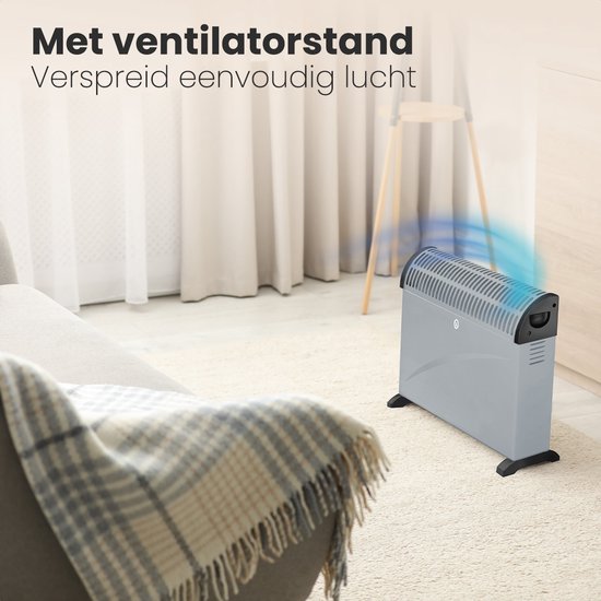 Grijze Auronic convectorkachel in een woonkamer, met blauwe luchtstromen die de ventilatorfunctie suggereren. Op een vloerkleed naast een fauteuil.