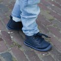 Kind met donkerblauwe leren veterschoenen met jeans op een bakstenen ondergrond.