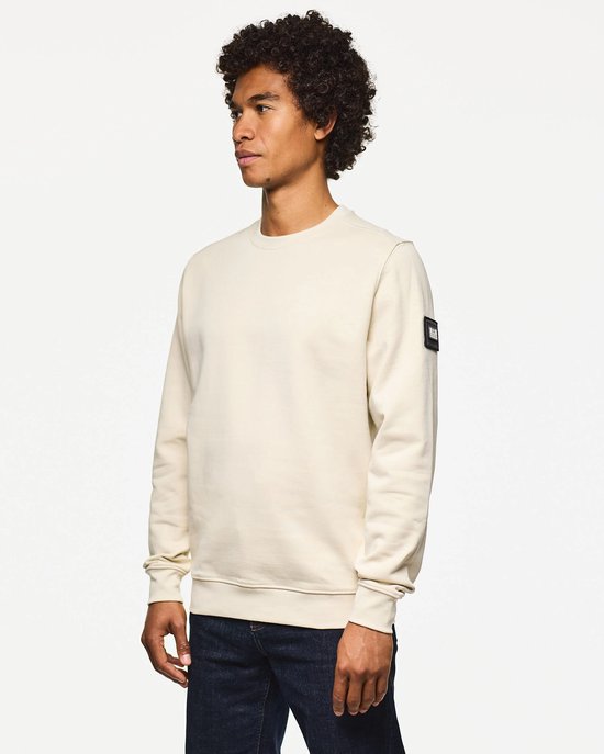Man met krullend haar, van opzij gezien, draagt een beige trui met een zwart label op de linker mouw.