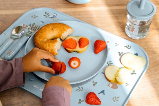 Assiette bleue pour enfant avec imprimé d'animaux et de champignons. Enfant coupant une fraise avec un petit couteau. Doigts d'enfant visibles.