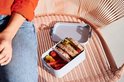 Boîte à pain grise ouverte avec sandwichs et légumes, posée sur une chaise.