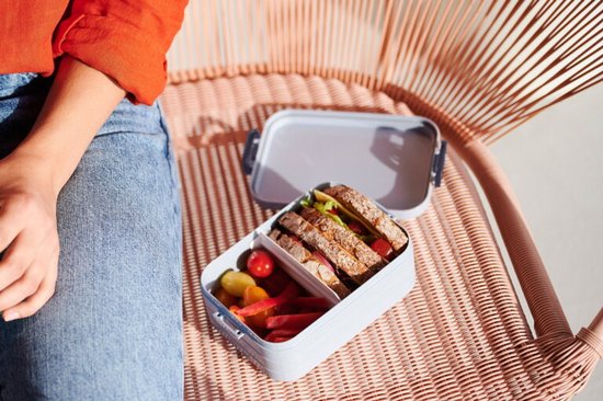 Boîte à pain grise ouverte avec sandwichs et légumes, posée sur une chaise.