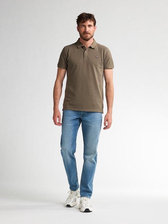 Man in een olijfgroen poloshirt, blauwe jeans en witte sneakers, volledig in beeld.