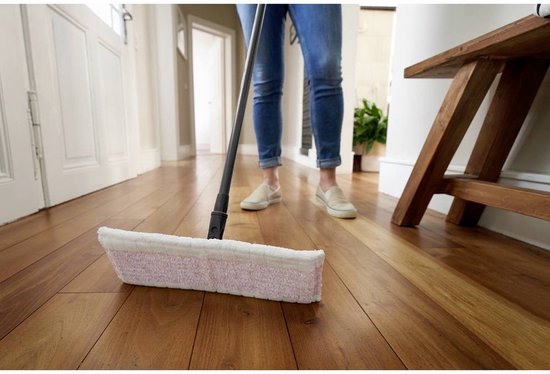 Une personne en jean et chaussures blanches passe une serpillière violette et blanche sur un parquet.