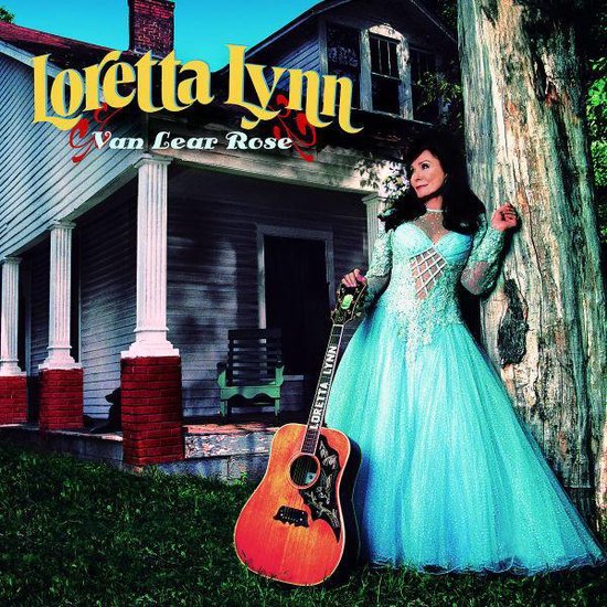 Loretta Lynn met gitaar bij huis, draagt een lichtblauwe jurk met glitter.