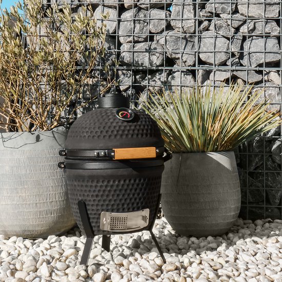 Zwarte kamado barbecue met houten handvat, staand op drie poten, in een tuin.