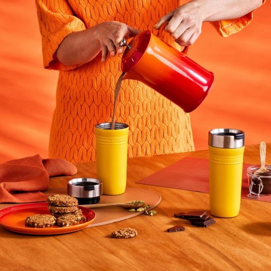 Een geel Le Creuset beker wordt gevuld met chocolademelk uit een rood-oranje thermoskan. Een tweede gele beker staat ernaast.