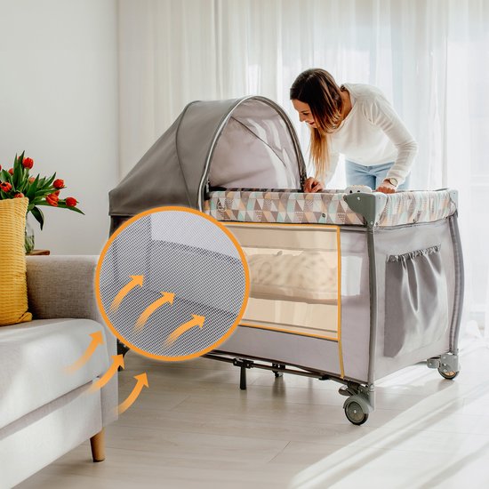 Vrouw kijkt naar een donkergrijs reisbedje met kap in een woonkamer.