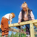 Twee meisjes op een houten speeltoestel, eentje met blond haar in een zwarte vest.