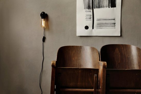 Edison-stijl lamp met gouden gloed aan een zwart snoer aan een muur naast twee houten stoelen en een abstract schilderij.