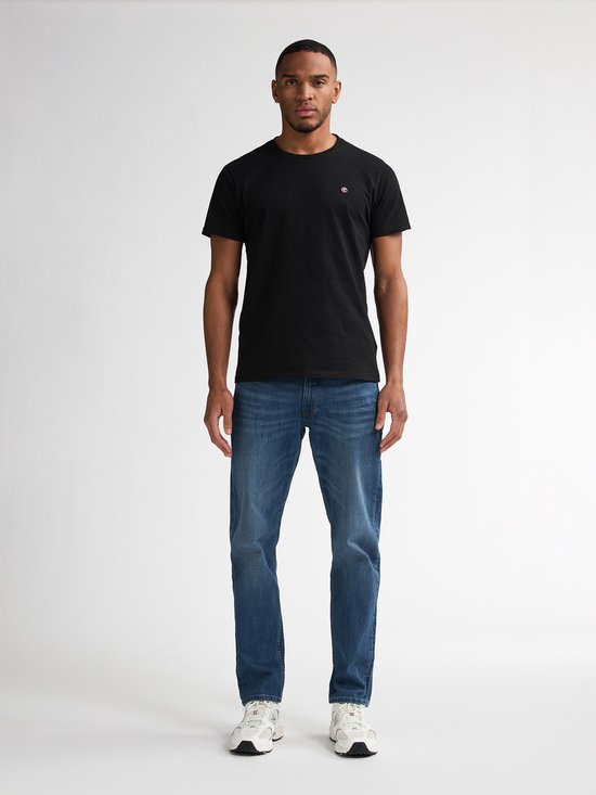 Man met de voorkant naar de kijker, draagt een zwarte t-shirt met klein logo en een blauwe jeans. Witte sneakers.