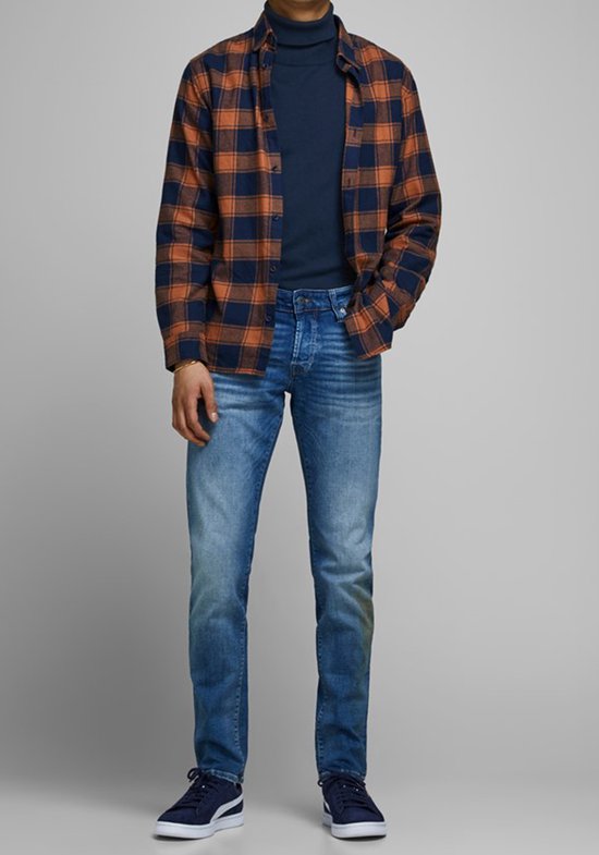 Man in een donkerblauwe coltrui, een openstaand geruit overhemd met oranje en marineblauwe ruiten, en een spijkerbroek.