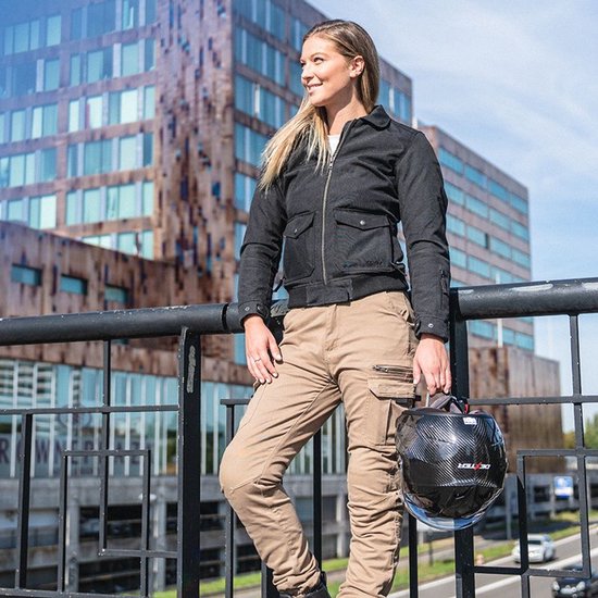 Vrouw in beige cargobroek en zwarte motorjas, buiten met helm in de hand.