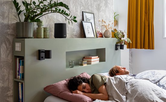 Zwarte Harman Kardon speaker boven een bed met slapend koppel.