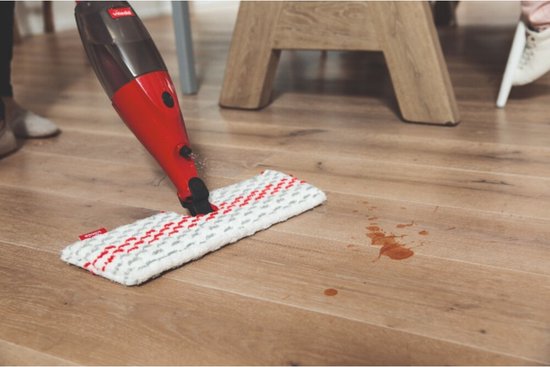 Le mop Vileda 1.2.Spray MAX nettoie une tache brune sur un plancher en bois. Le mop est blanc avec des carreaux rouge-gris.