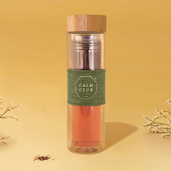 Glazen "Calm Club LDN" theefles met bamboedop, groen hoesje en oranje thee. Metalen infuser zichtbaar.