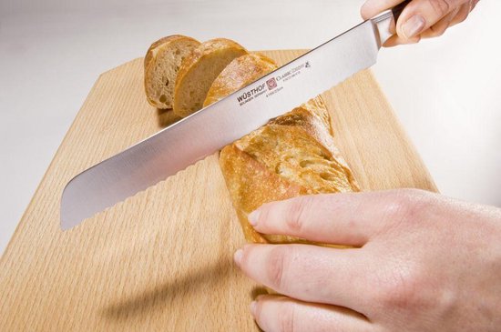 Een hand snijdt een brood met een Wüsthof broodmes op een houten snijplank.