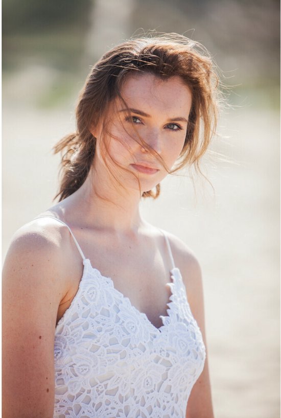 Model met bruin haar en sproeten draagt een witte kanten top, het haar wappert in de wind.