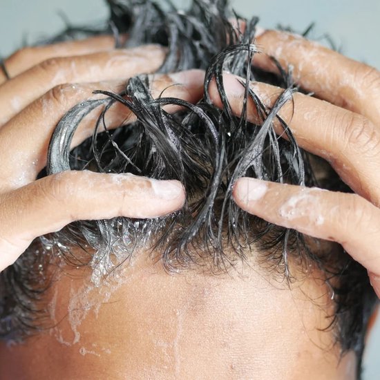 Detailopname van handen die donker haar met shampoo masseren.