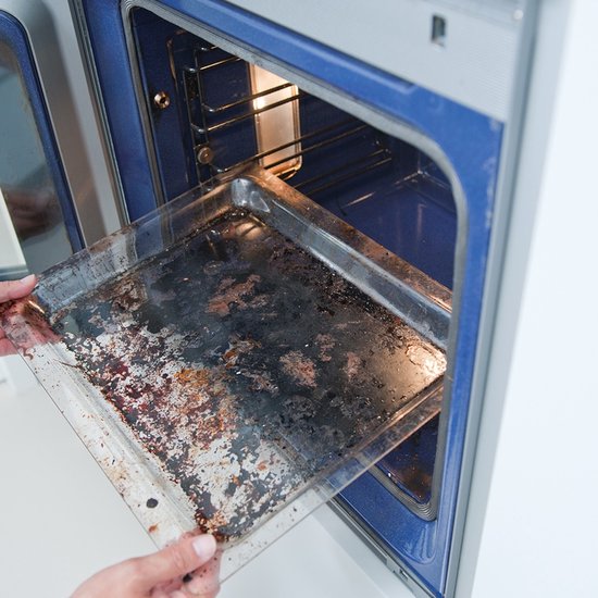 Vuile bakplaat wordt uit een oven gehaald. De bakplaat is zwart van het aangekoekte vet.