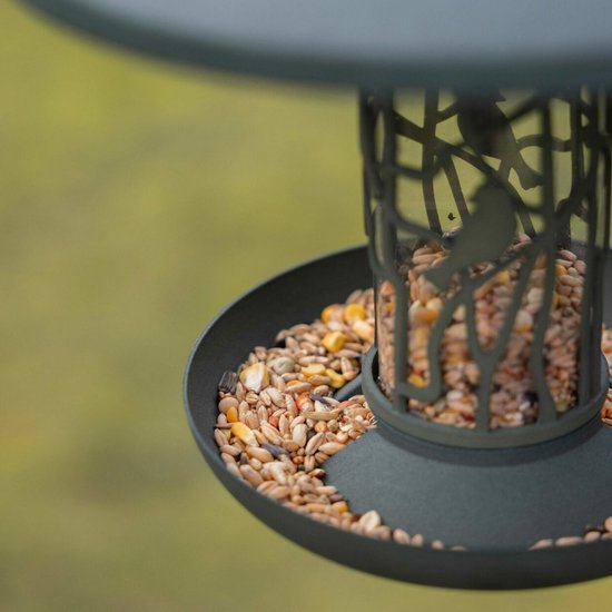 Close-up van de schotel van een groene vogelvoedersilo, gevuld met vogelzaad. De metalen decoratie toont takken en vogels.