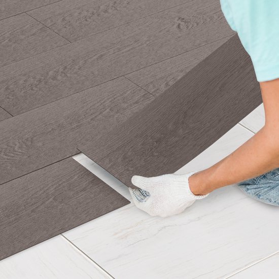 Een persoon met witte handschoenen legt een donkergrijze PVC-vloer met houtnerfpatroon.
