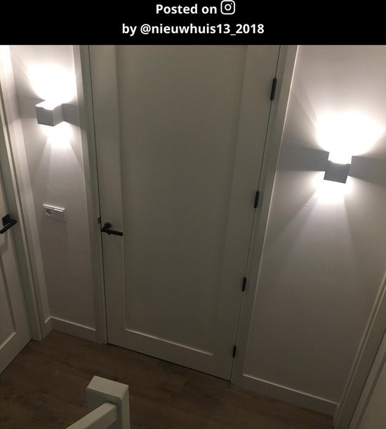 Twee witte wandlampen aan weerszijden van een witte deur, verlichten de gang met warm licht.