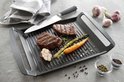 Gegoten aluminium grillplaat met ribbels, waarop twee steaks, een halve knoflook, asperges en wortel liggen. Peperkorrels en een mes liggen ernaast.