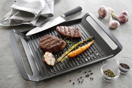 Gegoten aluminium grillplaat met ribbels, waarop twee steaks, een halve knoflook, asperges en wortel liggen. Peperkorrels en een mes liggen ernaast.