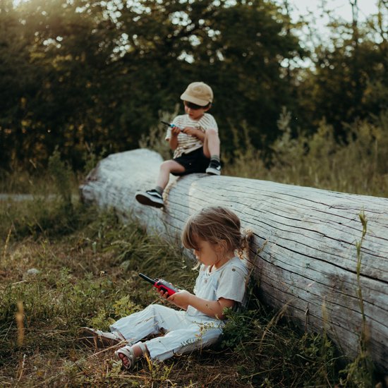 Twee kinderen met Kidywolf KIDYTALK walkietalkies op een boomstam en grasveld.