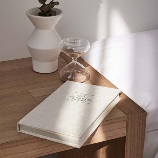 Linnenkleurige 'The Five Minute Journal' op een houten nachtkastje, naast een witte vaas en zandloper.