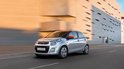 Zilverkleurige Citroën C1 rijdt op een straat met moderne architectuur op de achtergrond.