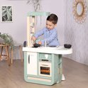 Een jongetje speelt met een mintgroene Smoby speelgoedkeuken in een woonkamer. De keuken heeft een koelkast, oven, gootsteen en kookplaat.