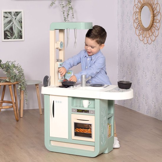 Een jongetje speelt met een mintgroene Smoby speelgoedkeuken in een woonkamer. De keuken heeft een koelkast, oven, gootsteen en kookplaat.