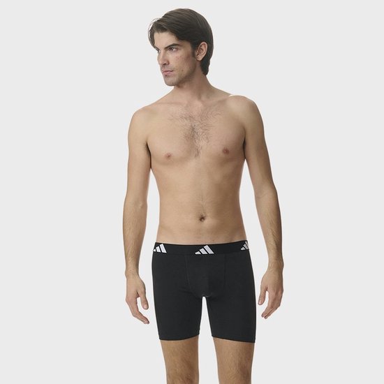 Man, volledig gekleed in zwarte Adidas boxershorts met witte logo's op de tailleband.