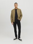 Een man staat met een beige bomberjack over een zwarte trui en zwarte jeans.