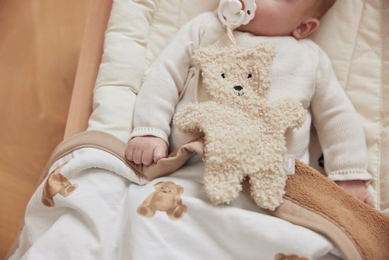 Bébé avec sucette et une peluche ours beige sur une couverture blanche à imprimé ours en peluche et bordure marron.
