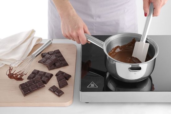 Een hand smelt chocolade in een pannetje op een kookplaat, met repen chocolade ernaast.
