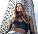 Een vrouw met lang bruin haar, die een zwarte top draagt, kijkt omhoog tegen de achtergrond van een hoog gebouw.