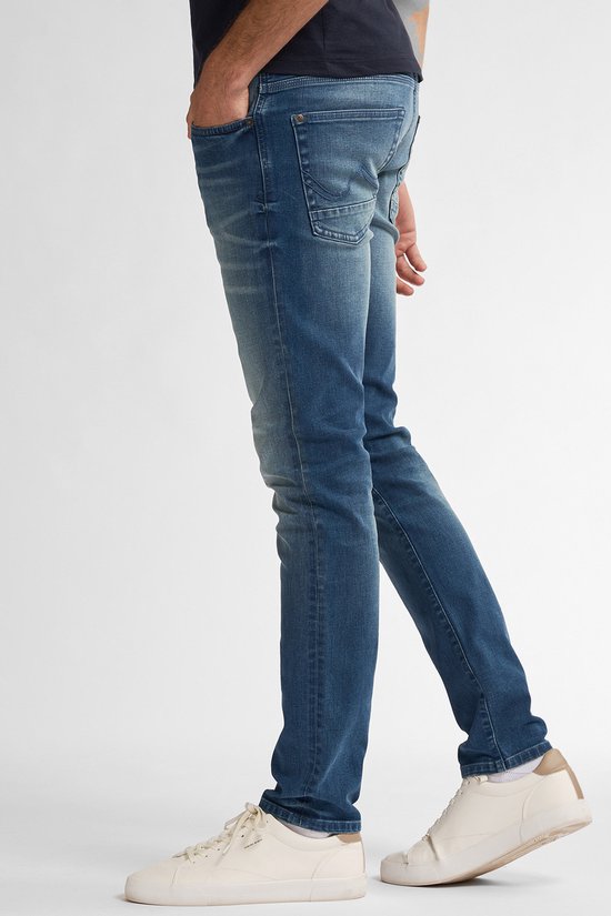 Man in een slim fit spijkerbroek, vaal blauw, met een marineblauw t-shirt. Van de zijkant gezien.