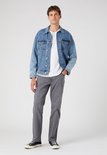 Man staat vol in beeld in een grijze jeans, wit t-shirt en een blauwe denim jas. Hij draagt sneakers.