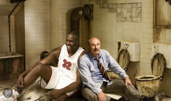 Shaquille O'Neal in basketbaltenue en Dr. Phil zitten vastgeketend in een vuile ruimte.