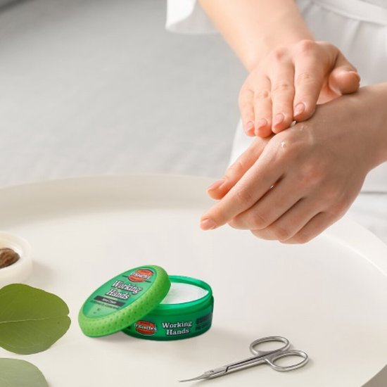 Een vrouw brengt O'Keeffe's Working Hands Handcrème aan op haar handen. Naast de groene pot ligt een schaartje.