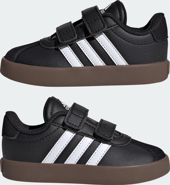 Twee zwarte Adidas kindersneakers met witte strepen en klittenbandsluiting, op bruine zolen.